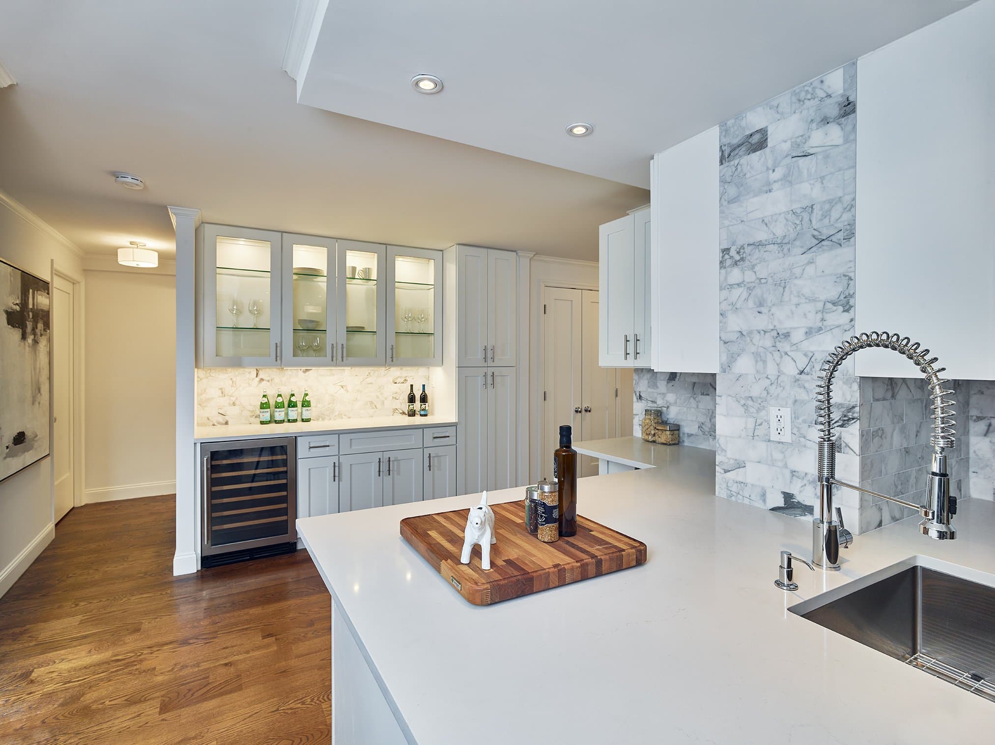 A wet bar with glass-front cabinets and a Carrara marble accent wall in the Greenbaum Residence